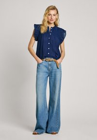 Blusa blu navy con maniche a volant, parte anteriore con bottoni, abbinata a jeans a gamba larga azzurri e una cintura marrone. Un look casual e texturizzato.