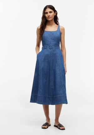 Kvinde med langt mørkt hår iført en ærmeløs, bælte denim midi kjole og sorte slides sandaler, stående mod en ensfarvet hvid baggrund.