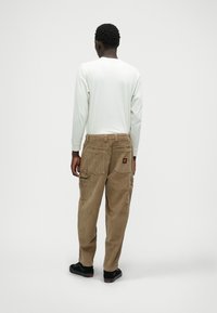 Καφέ καπρι παντελόνι από κορντούρα με πολλά τσέπες, φορεμένο με ένα ανοιχτόχρωμο μακρυμάνικο πουκάμισο. Μαύρα αθλητικά παπούτσια ορατά στα πόδια.