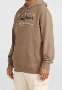 Sweat à capuche beige avec poche avant et cordons, arborant le texte "O'Neill California Santa Cruz The Original Surf Supply Co." en polices mixtes.