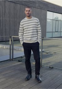 Ljust grå randig tröja, mörka byxor och svarta ankellångstövlar. Ståendes pose på en trädeck nära en modern byggnad med glasfönster.