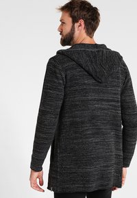 Cardigan negro de punto con un patrón texturizado, que presenta una capucha, mangas largas y bolsillos laterales. Ajuste holgado con un diseño más largo en la parte posterior.