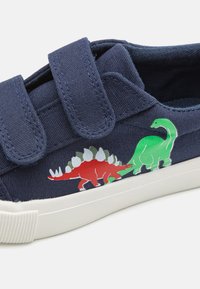 Chaussure en toile bleu marine avec deux straps en velcro. Présente des graphismes de dinosaures rouges et verts sur le côté. Semelle en caoutchouc blanche avec un motif texturé.