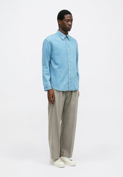 Homem com uma camisa de botão azul clara, calças largas cinzentas e sapatilhas brancas, posando contra um fundo branco simples.