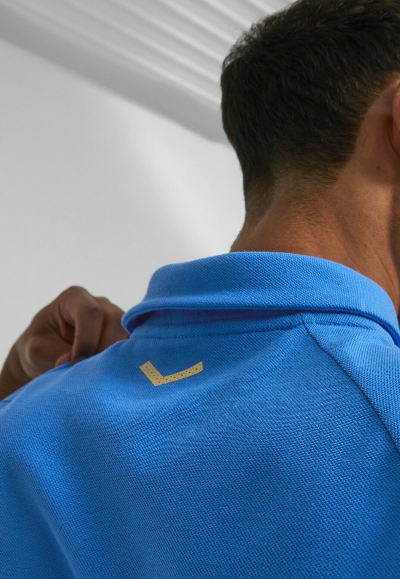 Homme portant un polo bleu avec un petit logo beige en forme de chevron dans le dos près du col, ajustant la chemise de la main.
