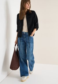 Kobieta w czarnej kurtce, beżowym topie, szerokich niebieskich dżinsach i białych tenisówkach, trzymająca dużą brązową torebkę, stojąca na tle jasnej ściany.