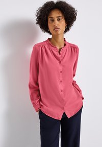 Blusa rosa a maniche lunghe con colletto arricciato, design con bottoni sul davanti e polsini arricciati, abbinata a pantaloni blu scuro. Tessuto di trama liscia.