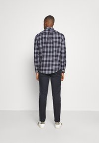 Man standing facing away wearing a navy, white, and brown plaid shirt, dark pants, and white sneakers with beige details.