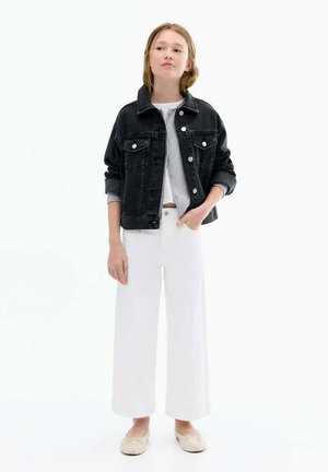 Giacca in denim nera con tasche a soffietto, chiusura a bottoni, camicia grigia sotto, pantaloni bianchi a gamba larga e scarpe slip-on chiare.
