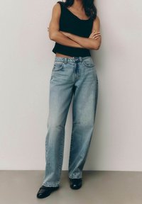 Jeans de corte ancho azul claro con un acabado desgastado, que cuentan con cinco bolsillos y presillas para el cinturón, combinados con una blusa sin mangas ajustada negra.