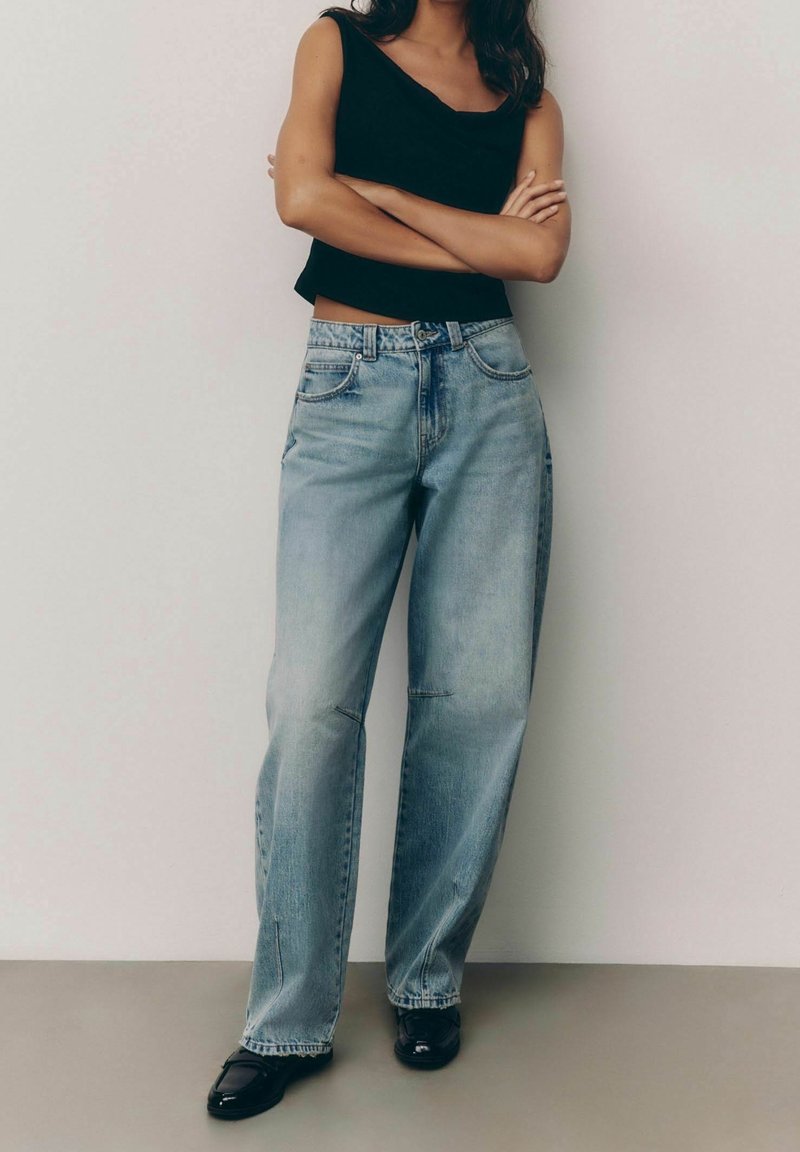 Jeans de corte ancho azul claro con un acabado desgastado, que cuentan con cinco bolsillos y presillas para el cinturón, combinados con una blusa sin mangas ajustada negra.