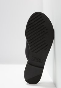 Semelle de chaussure noire avec des rainures horizontales et le logo "Tommy Jeans" embossé près du talon, positionnée verticalement contre un mur blanc.
