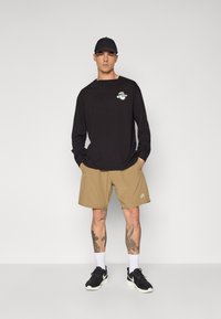 Black long-sleeve shirt with graphic logo, beige shorts, and black sneakers. The outfit features relaxed fit and cotton blend material.
