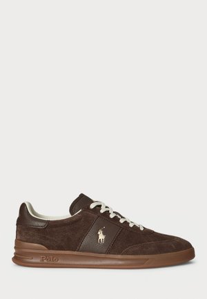 Brown suede sneakers featuring a tan rubber sole, flat white laces, and a gold logo accent on the side. Clean, casual design.