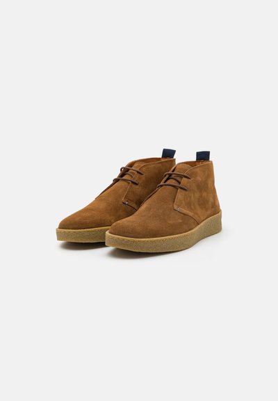 Braune Wildleder-Stiefeletten mit glatter Textur, ausgestattet mit einer Schnürung, Gummisohle und einem blauen Zugschlaufenakzent auf der Rückseite.