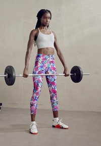 Športovec drží činku v vzorovaných, pestrofarebných legínach a svetlom crop topu, s bielymi a oranžovými športovými topánkami na neutrálnom pozadí.