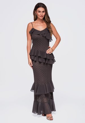 Femme portant une robe en dentelle noire sans manches avec des volants superposés et des fines bretelles, debout avec une main sur la hanche devant un fond uni.