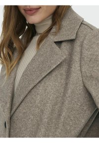 Femme portant un manteau en laine beige par-dessus un pull à col roulé léger à côtes, avec des cheveux lâches de couleur brun clair visibles.