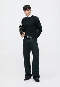 Svart ribbad tröja, mörk denim vida byxor, svart bälte med öglor, håller en svart kuvertväska, polerade svarta skor, minimal bakgrund.