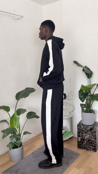 Camisola preta com riscas brancas, calças pretas a condizer com riscas laterais brancas, usadas com sapatos pretos. Ambiente interior com plantas em vasos.