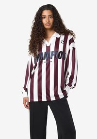 Vrouw met lang krullend haar, gekleed in een langemouw maroon en wit gestreept shirt met de tekst "CHAMPION" en een logo van een voetbal, in combinatie met zwarte broek.