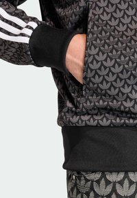 Čierna športová bunda s celoplošným vzorom lístkov, bielymi pruhmi na rukávoch a čiernymi rebrovanými manžetami. Na boku je viditeľná kapsa.