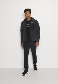 Giacca in pile grigia con colletto zip, dettagli neri e tasca con zip. T-shirt nera sotto con testo bianco. Pantaloni neri e scarpe da ginnastica.