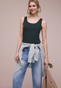 Jeune femme portant un débardeur vert foncé, un jean bleu clair, une chemise rayée nouée autour de la taille, tenant un sac à main beige sur un fond uni.