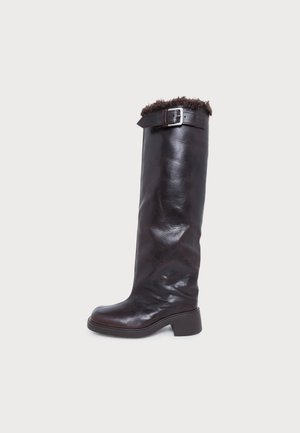 Botte haute en cuir marron foncé, à talon bloc bas, avec une bordure en fourrure en haut et une bride à boucle, sur un fond uni.