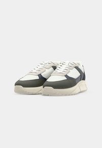 Sneakers in white, grey, and navy with a textured upper, padded collar, and thick rubber sole; features a lace-up front and decorative accents.
