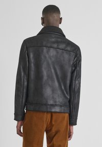 Man in een zwart schapenvacht jack en een paarse corduroy broek die met zijn rug naar de camera staat, tegen een effen achtergrond.