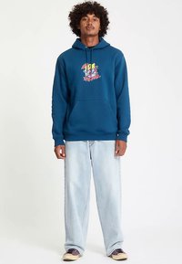 Blauer Baumwoll-Hoodie mit einem grafischen Motiv auf der Vorderseite und einer Kängurutasche, kombiniert mit hellblauen, locker sitzenden Jeans und dunklen Schuhen.