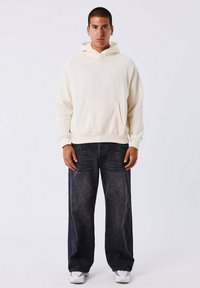 Jonge man met kort haar in een crèmekleurige hoodie, losse zwarte jeans en witte sneakers, staand tegen een effen lichte achtergrond.