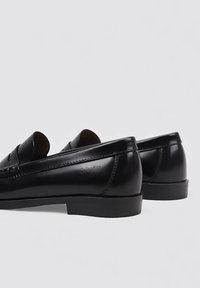 Mocasines negros de cuero con acabado liso, con una correa decorativa y detalle de costuras. Suela plana de goma con logotipo en relieve.