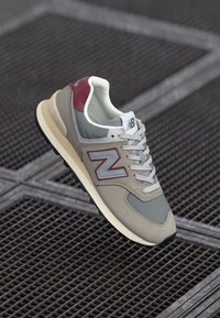 Sneaker grigia e beige con logo rosso e argento, caratterizzata da un upper testurizzato, collo imbottito e lacci bianchi su una suola nera.