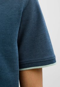 Dunkelblaues Poloshirt mit Kragen, der einen hellgrünen Rand hat, aus strukturiertem Stoff und mit kurzen Ärmeln. Nahaufnahme, die das Detail des Ärmels zeigt.