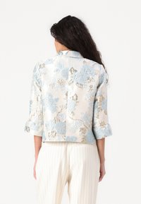Kukka jacquard-takki kerma- ja vaaleansinisessä värissä, jossa on metallinhohtoisia kultaisia yksityiskohtia, kolmanneksen mittaiset hihat ja lyhennetty malli.