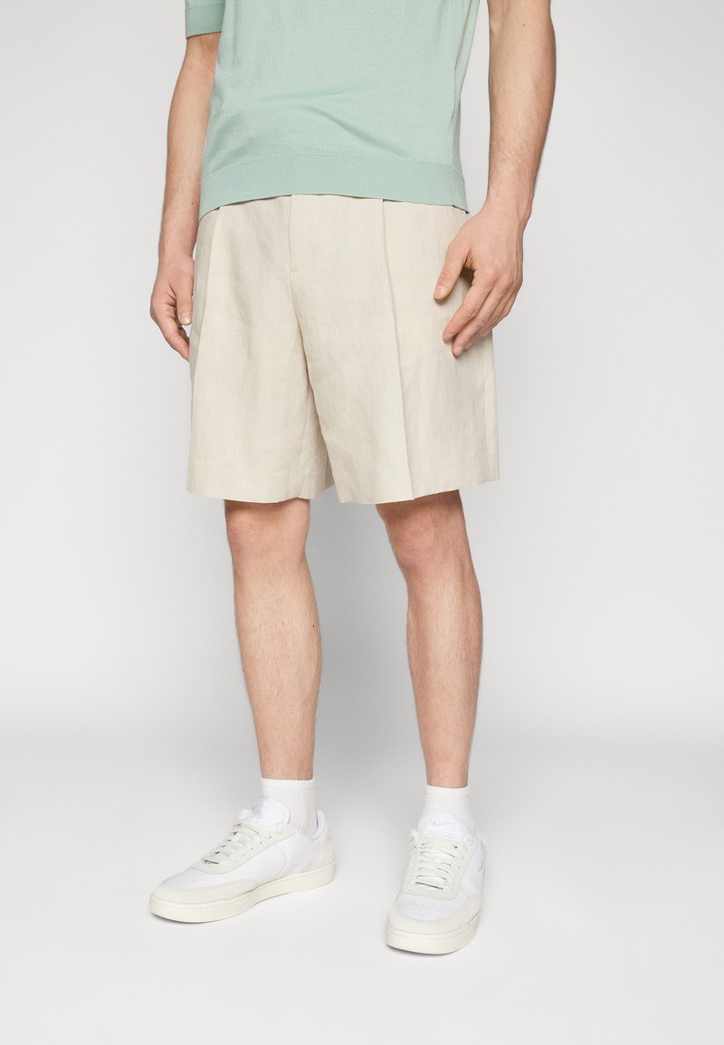 Shorts beiges ajustés avec des plis, en tissu léger. Associés à un haut en tricot vert clair et des baskets blanches.