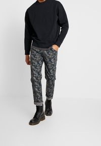 Sudadera negra con un acabado texturizado, combinada con pantalones de camuflaje en tonos grises y verdes, arremangados, complementados con botas negras.