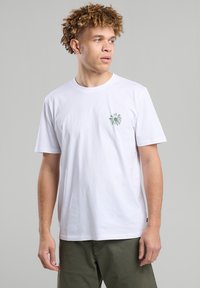 White cotton t-shirt featuring a green floral graphic on the left chest, short sleeves, and a relaxed fit design.