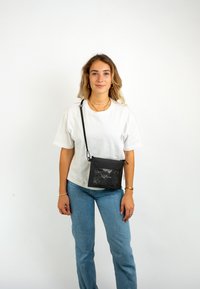 Fekete keresztpántos táska átlátszó elülső zsebbel, téglalap alakú, sima felülettel és állítható pánttal. Fehér ing fölött viselve.