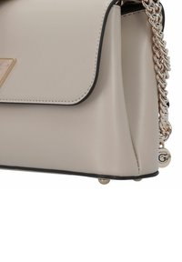 Beige leren handtas met een gouden kettingband, zwarte randen, driehoekig logo-detail en metalen voetjes voor stabiliteit.