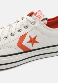 Zapatilla de lona blanca con estrella bordada naranja y diseño de chevrones. Presenta ojales metálicos, cordones planos y suela de goma negra.