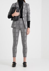 Femme portant un col roulé noir, un blazer double boutonnage à carreaux gris, un pantalon assorti à carreaux, une pochette en cuir noire et des mocassins noirs avec un détail doré.