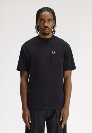 Homme aux cheveux tressés portant une chemise noire à manches courtes avec une poche poitrine ornée d'un petit logo de couronne de laurier blanche.