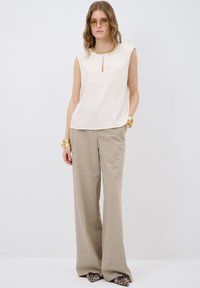 Top sin mangas color beige con escote en forma de ojal y cuello adornado, combinado con pantalones de pierna ancha marrón claro y zapatos con estampado.