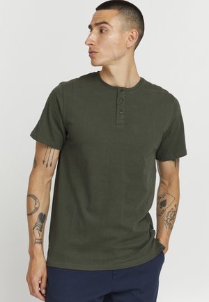Olive green short-sleeve t-shirt, crew neck with three buttons, soft cotton fabric, straight cut, visible tattoo on arm, worn with navy pants.