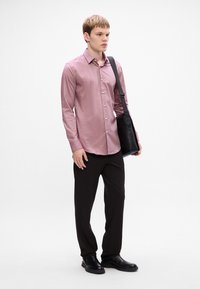 Camicia button-up rosa chiaro con una texture liscia, abbinata a pantaloni scuri e stivali neri. Una borsa nera a tracolla è indossata attraverso il corpo.