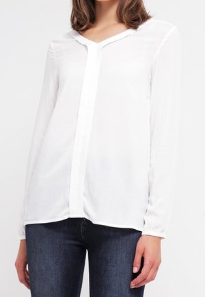 Kobieta ubrana w białą, długą bluzkę z ukrytym zapięciem na guziki oraz ciemnoniebieskie dżinsy, przedstawiona od szyi do bioder.