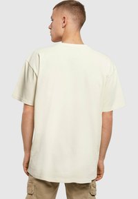 Tricou supradimensionat de culoare crem, cu mâneci scurte, decolteu rotund și textură netedă, văzut din spate pe un fundal neutru.